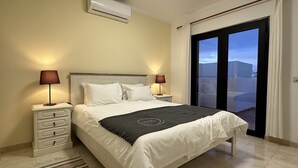 Ein Schlafzimmer mit einem Bett, zwei Nachttischen mit Lampen, einem weißen Beistelltisch, einem großen Fenster mit Aussicht und einer Klimaanlage an der Wand.