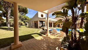 Ein überdachter Außenbereich mit Pool, Liegestühlen und einem Haus im Hintergrund.