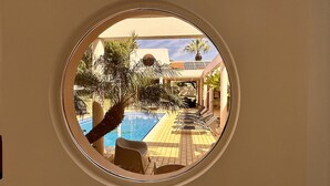 Blick durch ein rundes Fenster auf einen Poolbereich mit Liegestühlen und Palmen.
