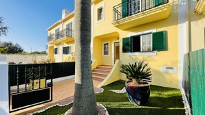 A two-story yellow building with green shutters, a gated entrance, and a small garden area with a palm tree and a large planter. - Mosqueira by Check-in Portugal (Albufeira)