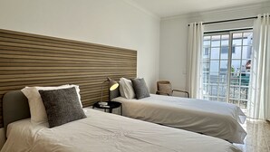 Ein Hotelzimmer mit zwei Betten, einem hölzernen Kopfteil, einem Nachttisch mit Lampe und Blick auf die Stadt durch ein Fenster mit weißen Vorhängen.