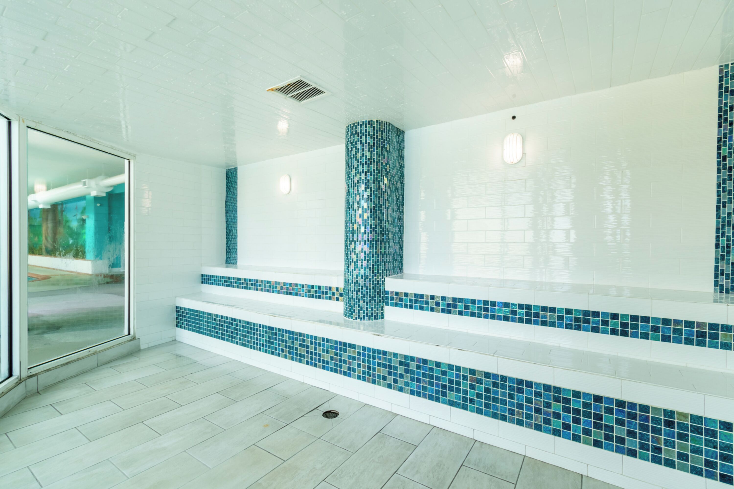 Un baño moderno con paredes de azulejos blancos, mosaicos azules en los escalones y una puerta de cristal que da a un área exterior.