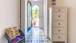A hallway with a patterned floor, a wicker chair with a striped cushion, a white dresser, and a door leading to an outdoor area with plants. - Capri Turquoise by Napoliapartments (Capri)