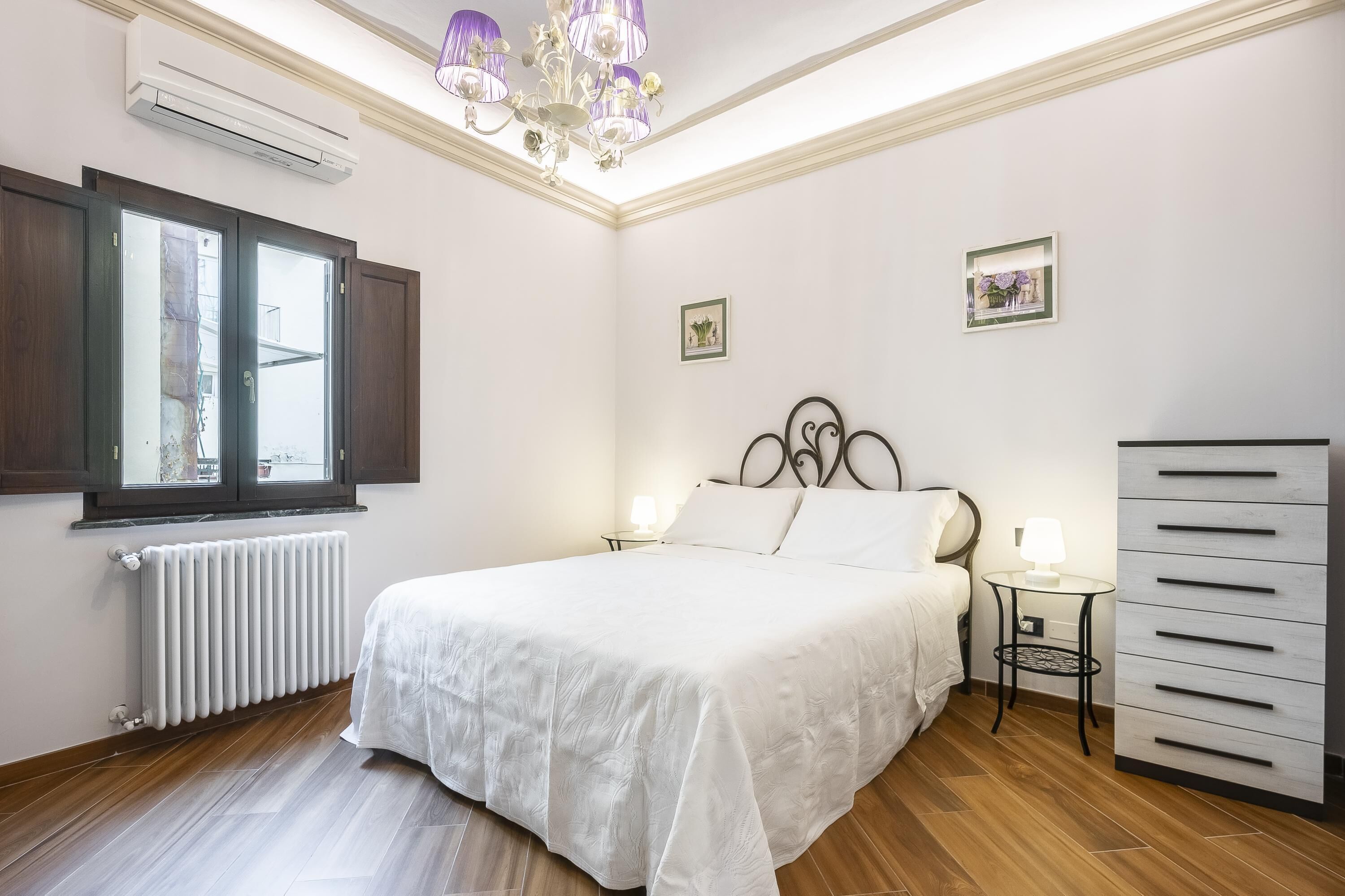 A bedroom with a bed, a radiator, a window, a chandelier, and a dresser.