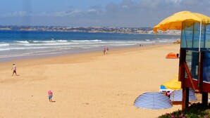 A beach with a lifeguard tower, umbrellas, and a clear view of the ocean and distant buildings. - Praia by Check-in Portugal (Albufeira)