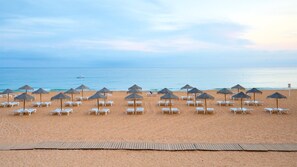 A beach with rows of sun loungers and thatched umbrellas, a wooden boardwalk, and the ocean in the background.