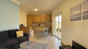 Un espacio habitable compacto con sofá, una mesa pequeña, una zona de cocina con armarios y una mesa de comedor, además de un ventanal corredero que da a un área exterior.