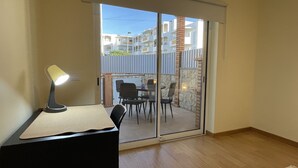 Una habitación con escritorio, silla y una mesa con lámpara. Hay una puerta corredera de vidrio que da a un espacio exterior con mesa y sillas.