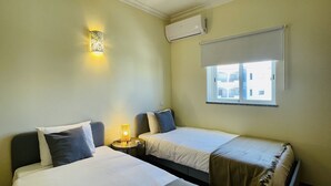 A hotel room with two beds, a bedside lamp, a window with blinds, and an air conditioning unit.