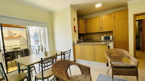 A compact kitchen with wooden cabinets, a microwave, and a coffee maker. There is a dining table with chairs and a wicker chair.