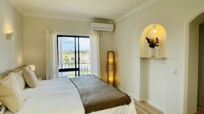 Une chambre à coucher comprenant un lit, une table de chevet, une étagère murale avec un vase, un appareil de climatisation et une vue sur l’extérieur par une fenêtre avec des rideaux.