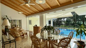 A spacious indoor area with a glass table surrounded by wicker chairs, a ceiling fan, and a view of a swimming pool outside. - Azaleas by Check-in Portugal (Vilamoura)