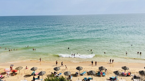 Uma praia com águas cristalinas, pessoas nadando e caminhando, e guarda-sóis.
