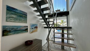 Uma escadaria moderna com grades de metal, que leva a um andar superior. Há três quadros com cenas costeiras na parede. Um cesto vermelho está sobre um banco de madeira, abaixo da escadaria.