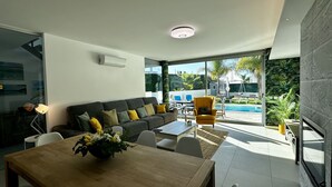 Uma sala de estar moderna com sofá, mesa de centro e cadeiras. Há uma mesa de jantar com um vaso de flores. Uma janela grande oferece vista para uma piscina e vegetação lá fora.