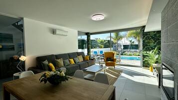 Un salon moderne avec un canapé, une table basse et des fauteuils. On y trouve également une table à manger ornée d’un vase de fleurs. Une grande fenêtre offre une vue sur une piscine et sur la verdure environnante.