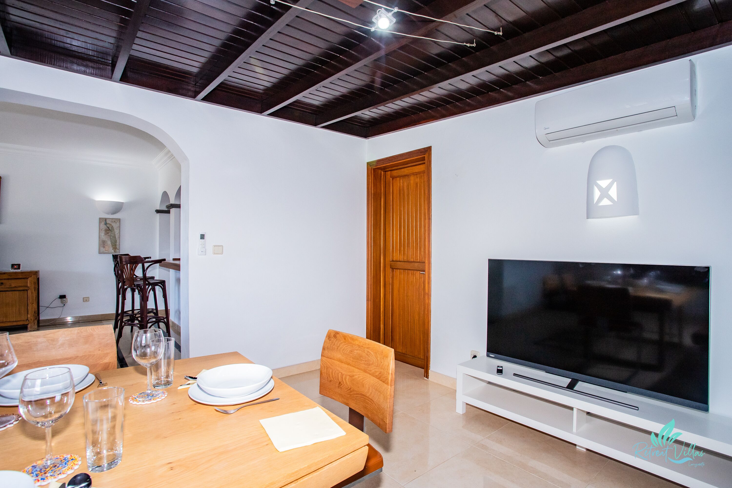 Un comedor con una mesa de madera dispuesta para cuatro personas, un televisor sobre un soporte blanco y una puerta de madera al fondo.