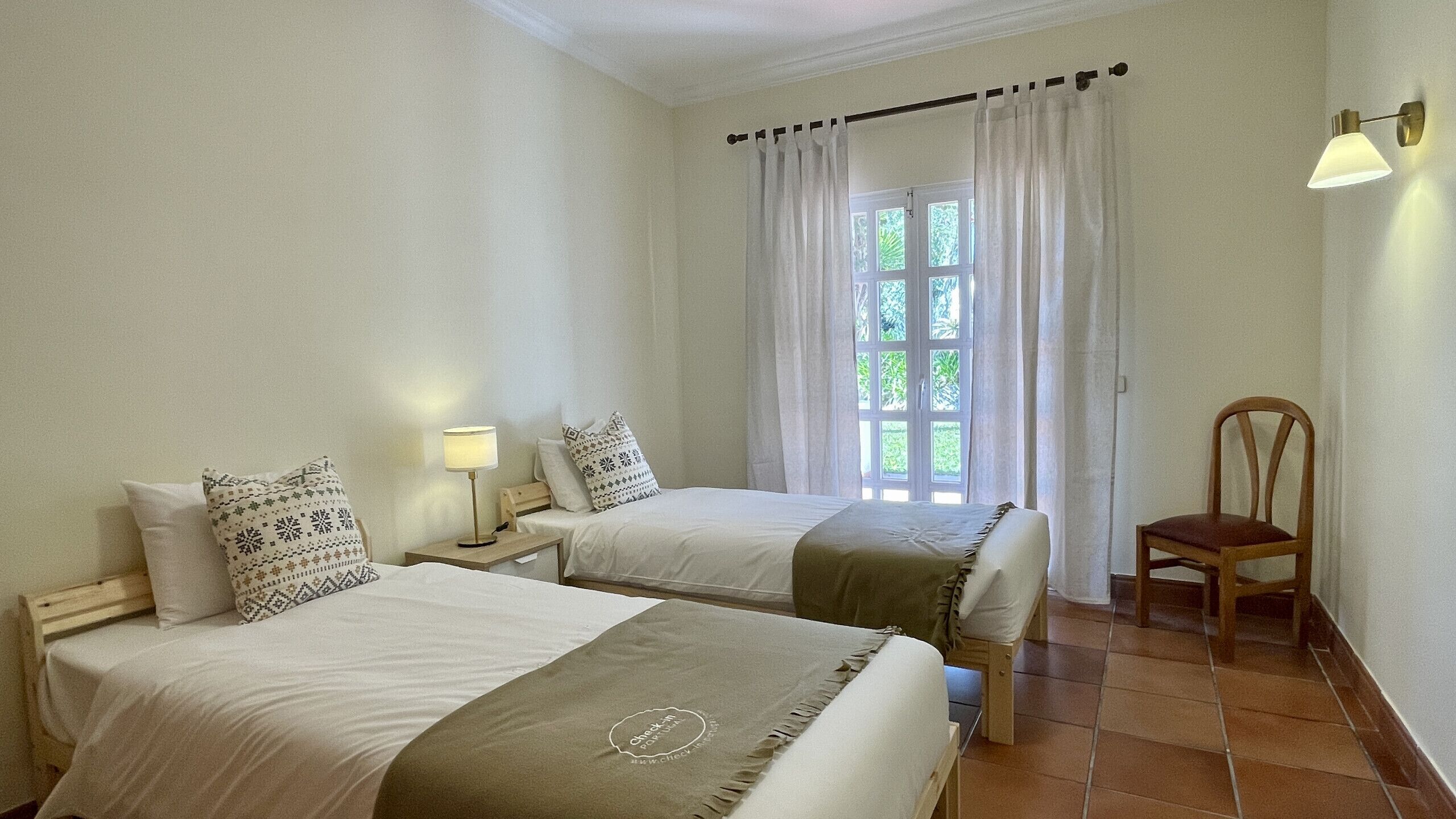 A hotel room with two beds, a chair, a window with curtains, and a wall-mounted lamp.