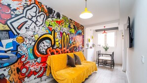 Un salon agrémenté d’une fresque murale colorée, avec un canapé jaune, une table à manger et des chaises, et un téléviseur fixé au mur.
