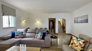 Ein Wohnzimmer mit einer Couch, Sesseln, einem Couchtisch und einem Essbereich mit Stühlen und einem Tisch. Eine Treppe führt zu einer weiteren Ebene.