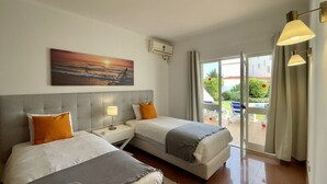 Ein Hotelzimmer mit zwei Betten, einem Sonnenuntergangsgemälde, einer Klimaanlage und Blick auf Pool und Garten durch die Glastür.