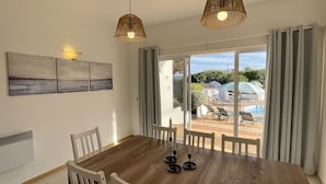A dining area with a wooden table and chairs, a white radiator, and a view of a pool and garden through large windows. - Nicola by Check-in Portugal (Albufeira)
