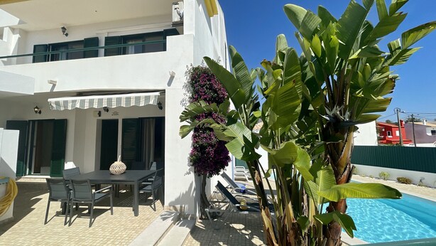 A white building with a pool, outdoor seating, and lush greenery.