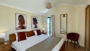 A hotel room with a large bed, a red chair, a nightstand with a lamp, and two framed artworks on the wall.