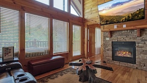 A cozy cabin interior with a stone fireplace, wooden floors, a leather sofa, and large windows with blinds. - Close to Pigeon Forge and Dollywood with mountain views (Sevierville)