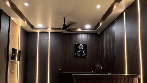 Ett välkomnande område med modern design, med en receptionsdisk som har en dekorativ lampa i form av en blomma, en takfläkt och en skylt med texten ”The Patel Stadium Hotel”.