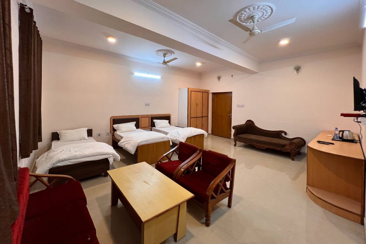 Habitación de hotel con dos camas individuales, una mesa de madera, un sillón rojo, un sofá, un ventilador de techo y una televisión.