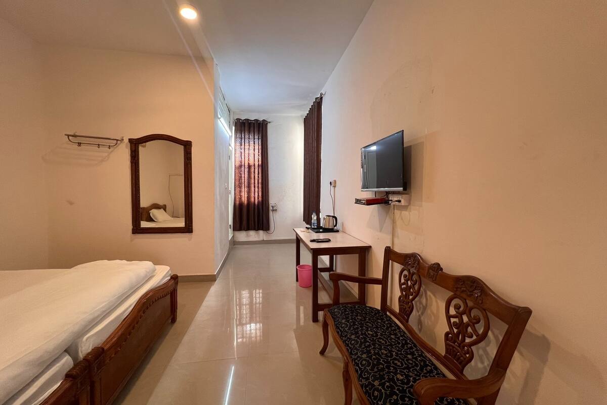 Una habitación de hotel con una cama, un banco de madera, un televisor fijado a la pared y una mesita con un hervidor eléctrico y un control remoto.