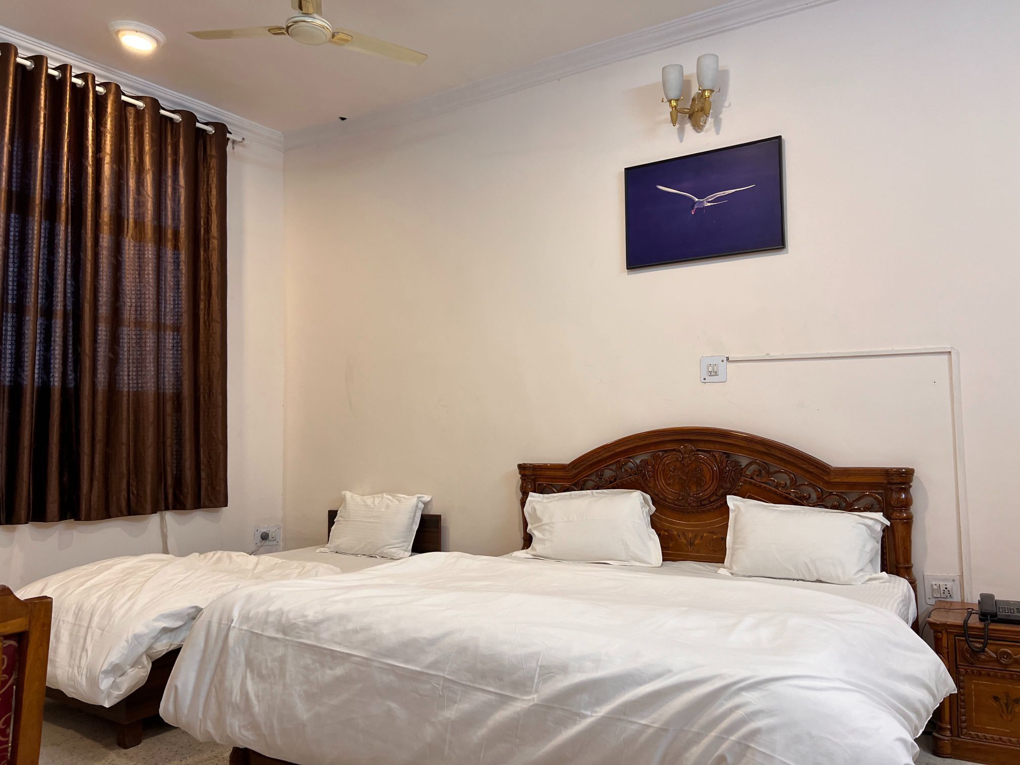 A hotel room with two single beds, a ceiling fan, a wall-mounted light fixture, a painting of a bird, and a bedside table with a phone.