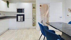 A modern kitchen with white cabinets, a microwave, an oven, and a wine bottle on the counter. There is a dining area with a marble table and blue chairs. - Cosy and Luxurious House for 10 Guests in Darwen (Darwen)