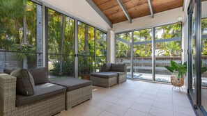 Un espace extérieur aménagé, abrité et doté de mobilier en osier, avec de grandes fenêtres offrant une vue sur la végétation tropicale.