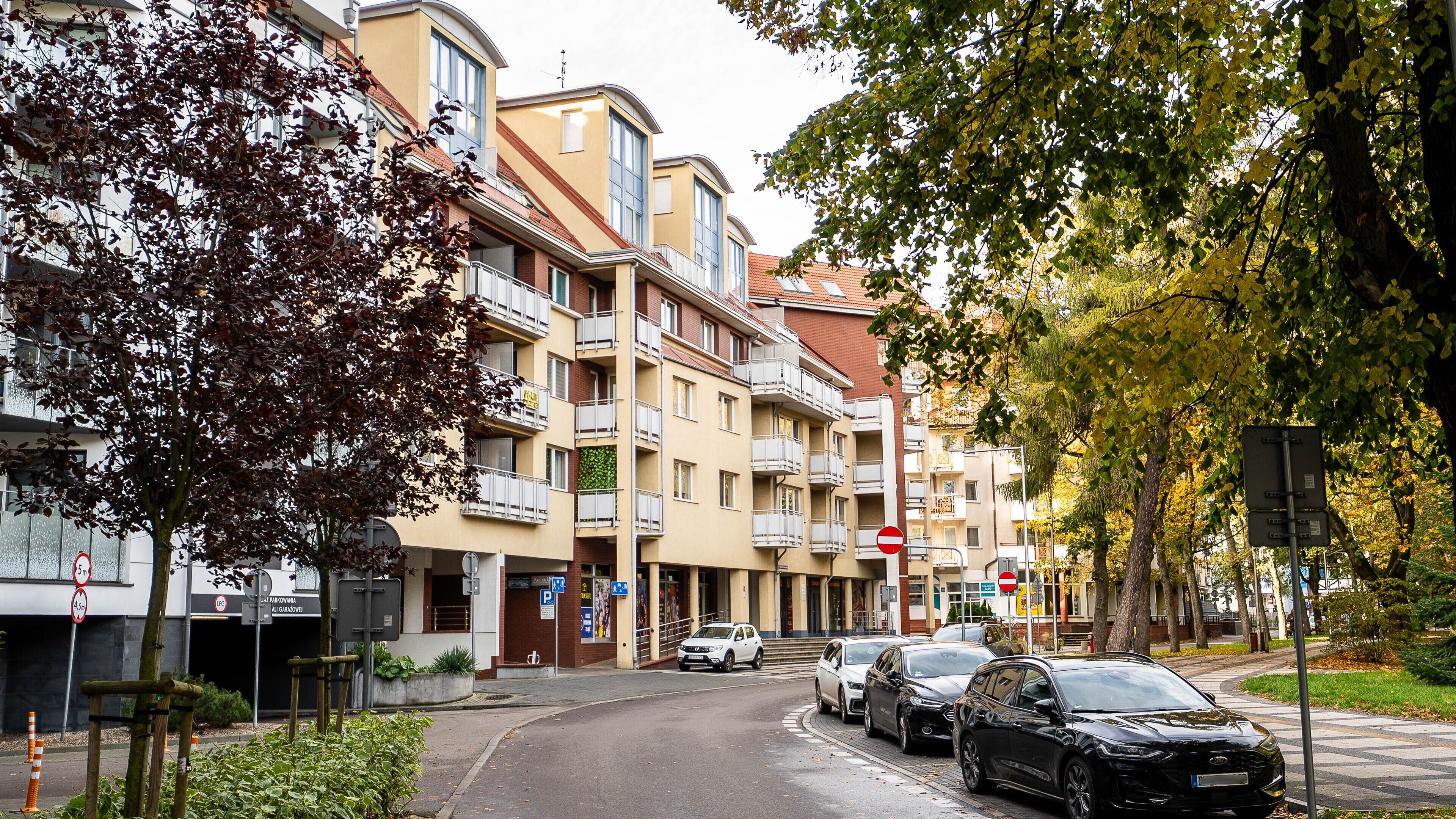 A multi-story residential building with balconies, a parking area, and a street with cars.