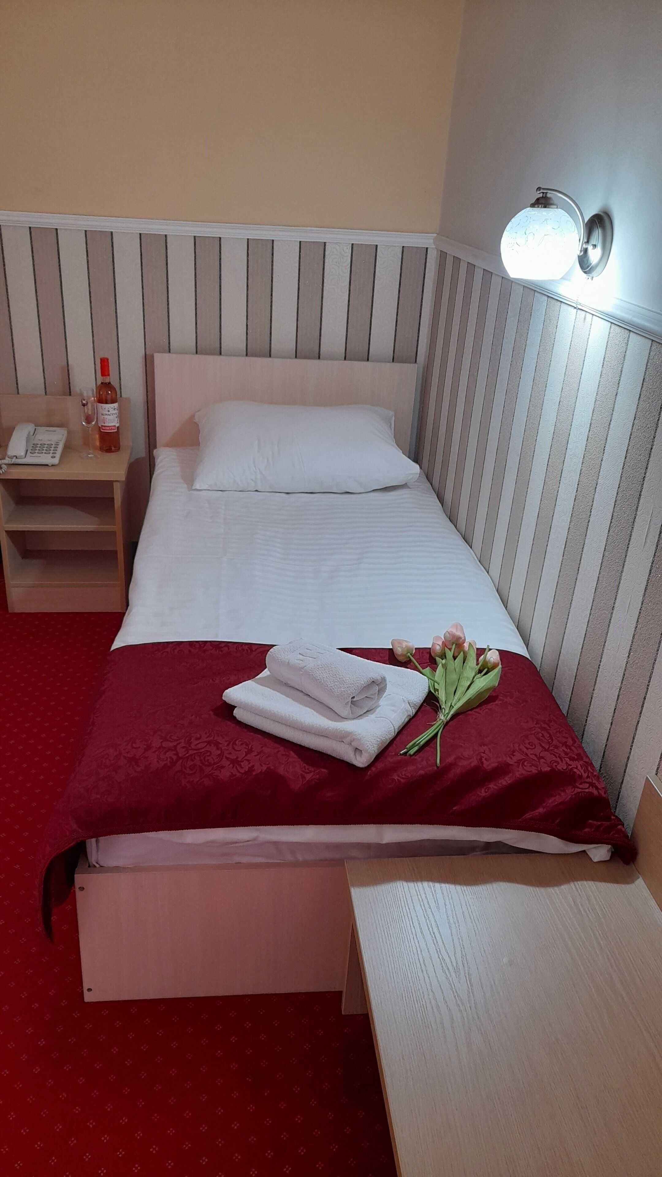 A neatly made bed with white linens, a red throw blanket, and a bouquet of flowers. There is a bedside table with a lamp, a phone, and a bottle of water.