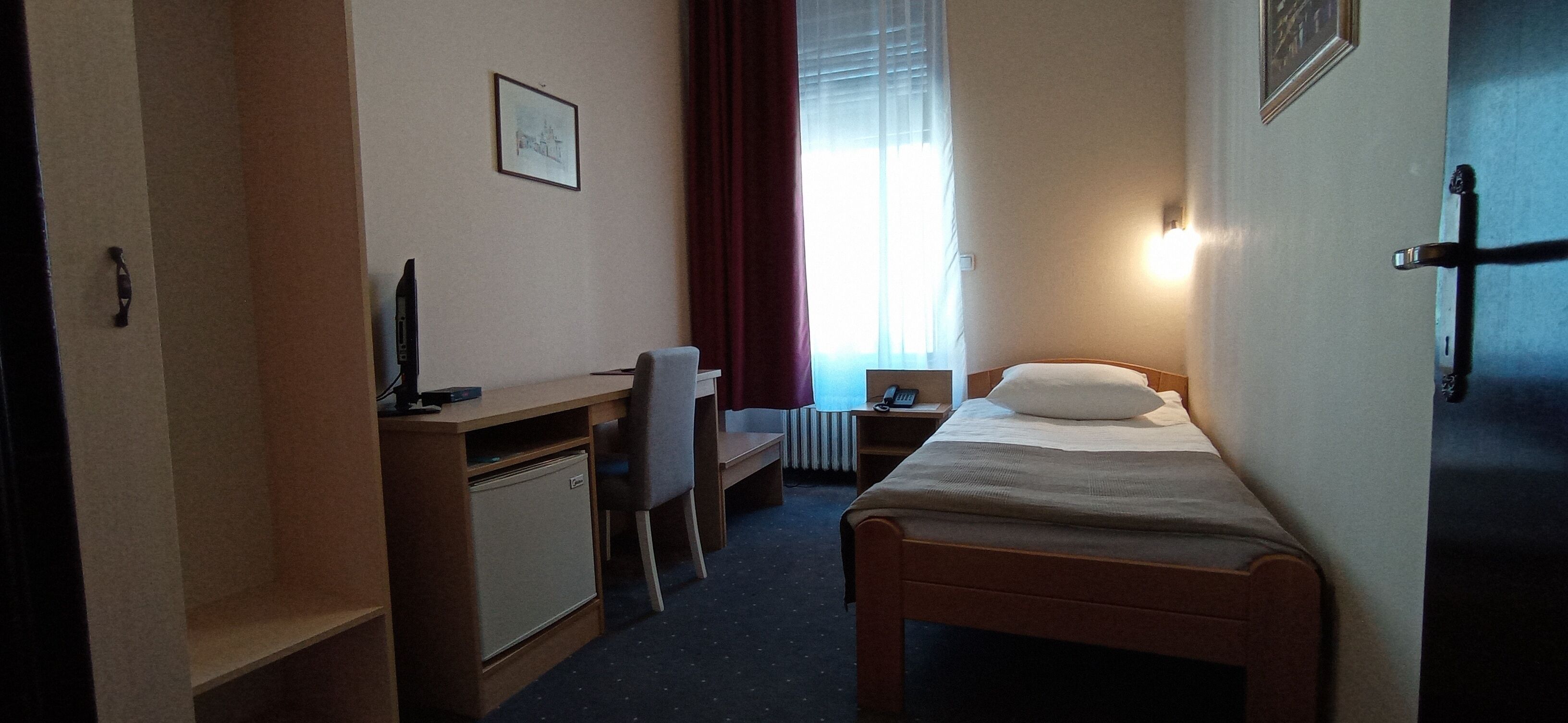 A hotel room with a single bed, a desk with a chair, a small refrigerator, and a window with curtains.