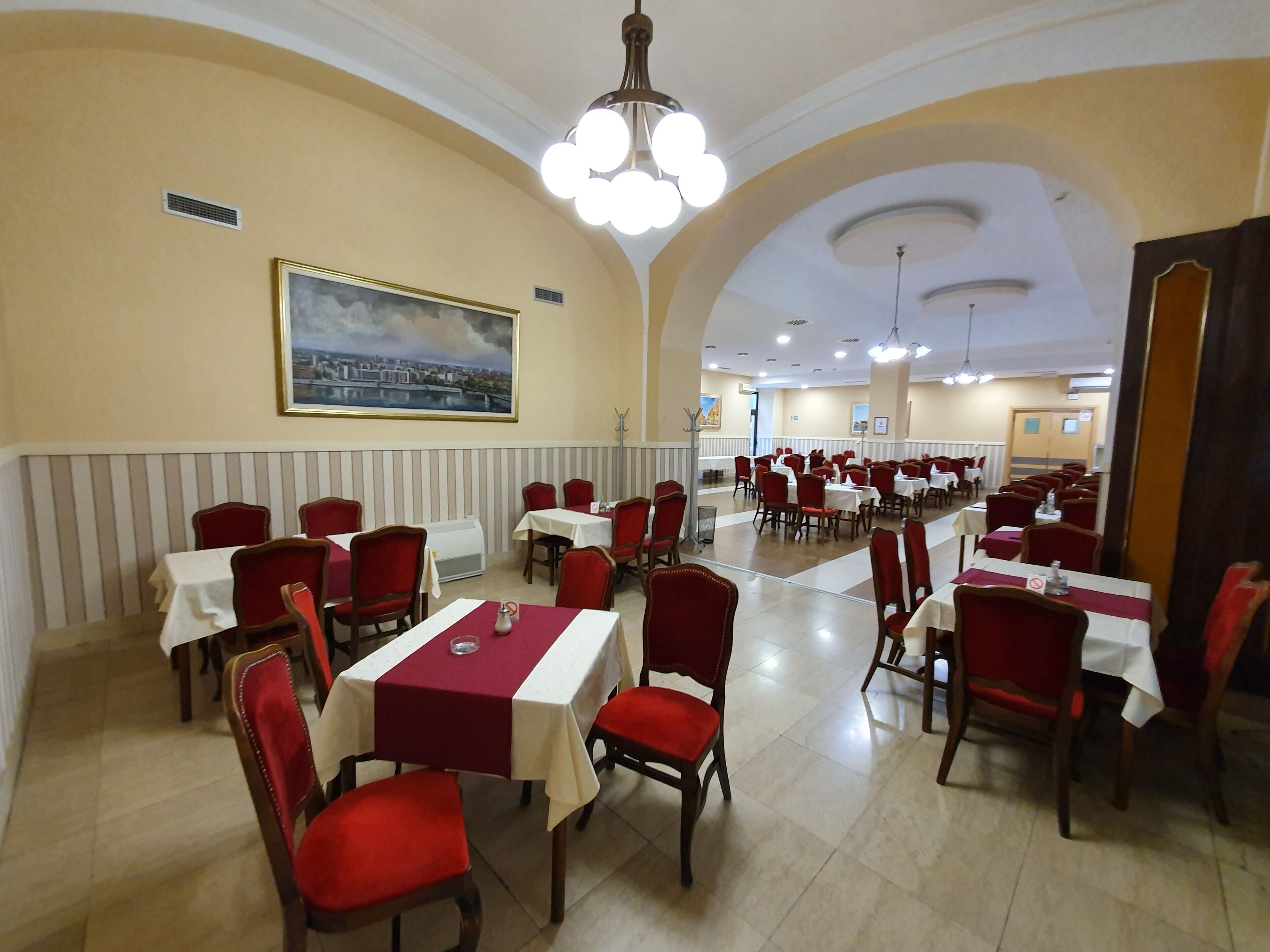 A spacious dining area with multiple tables and red chairs, featuring a large cityscape painting on the wall and a high ceiling with chandeliers.