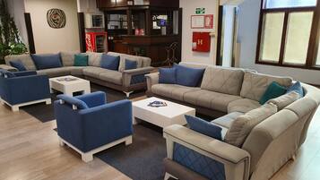 A modern lounge area with sofas, armchairs, and a coffee table. There is a vending machine, a fire extinguisher, and a window with a view.