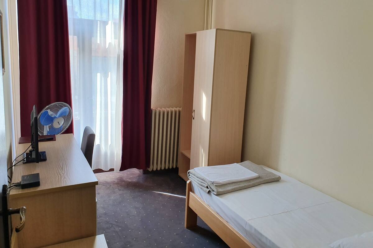 A small, tidy room with a bed, a desk with a computer, a standing fan, and a radiator.