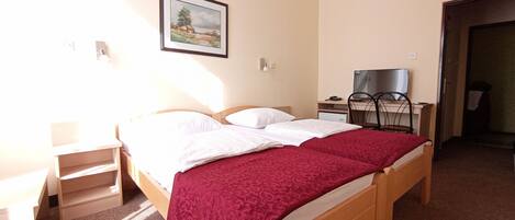 A double bed with white and red bedding, a desk with a chair, a television, and a framed picture on the wall.