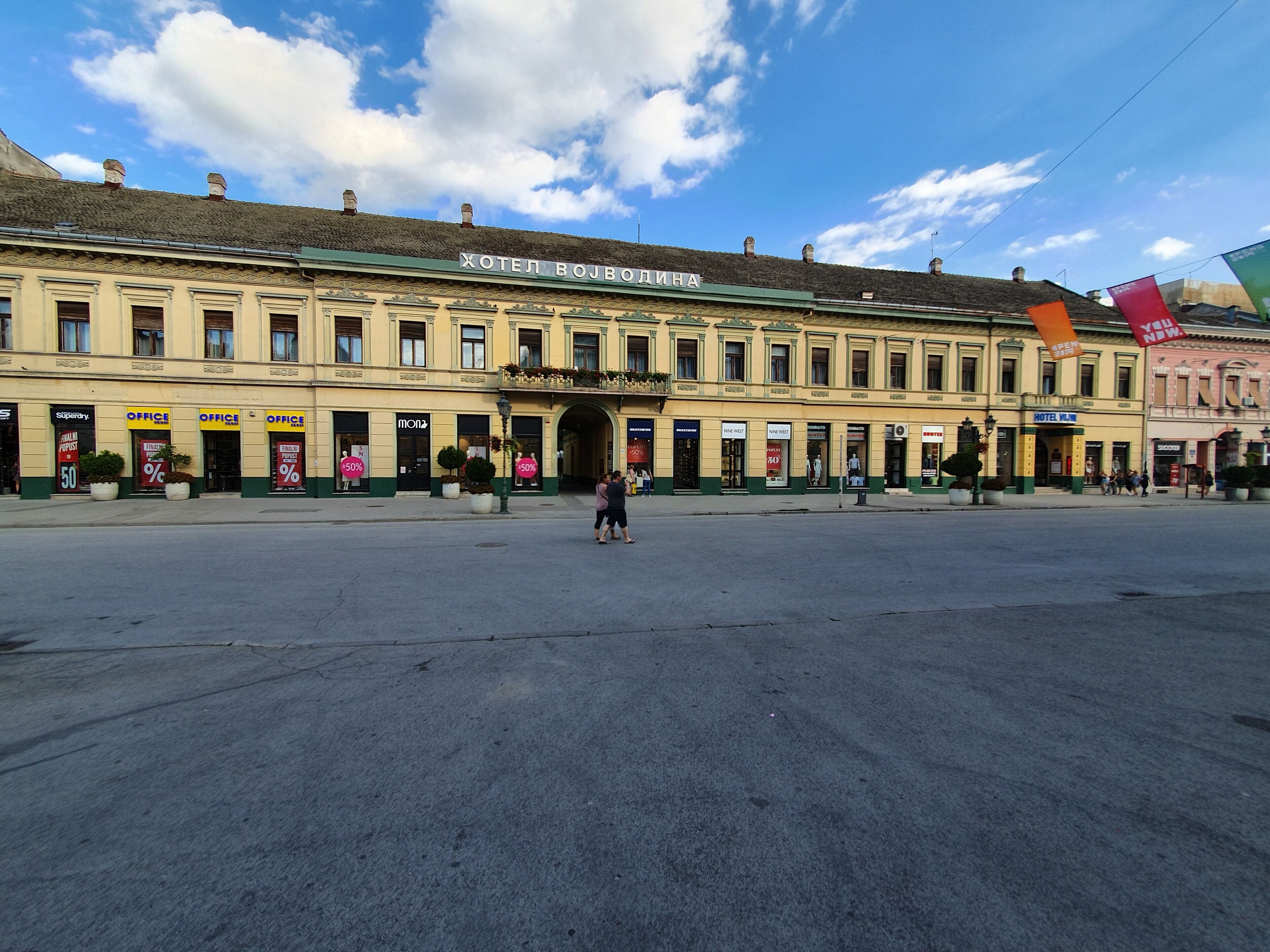 A historic building with a central archway, multiple storefronts on the ground floor, and a clear sky above.
