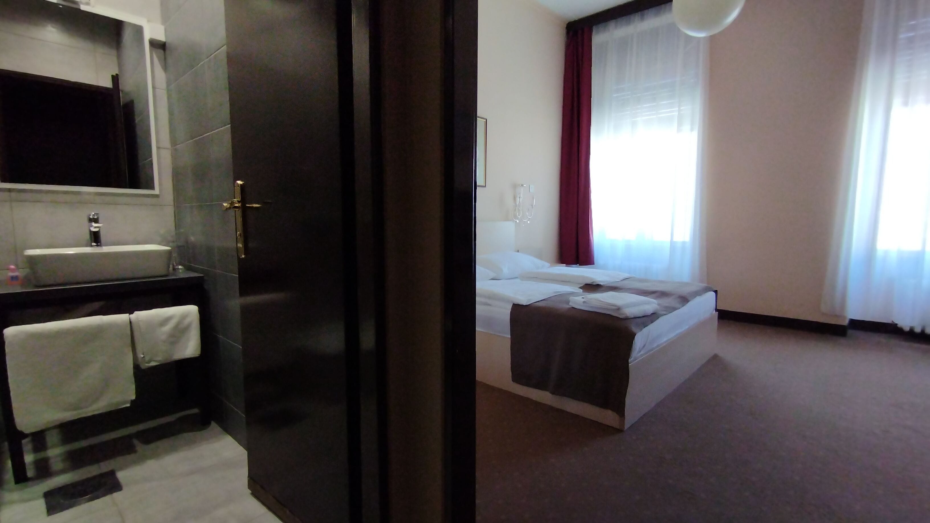 Habitación de hotel con baño que incluye un lavamanos, espejo y toallero. La habitación tiene una cama con edredón blanco y una manta marrón.