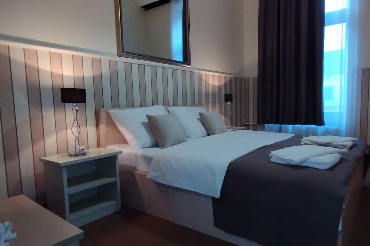 A hotel room with a large bed, bedside tables, a nightstand, a mirror, and a window with curtains.