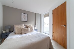 A bedroom with a large bed, a bedside table with lamps, a framed picture on the wall, and a wooden door. - 3 Bed Flat by Limehouse Basin (London)