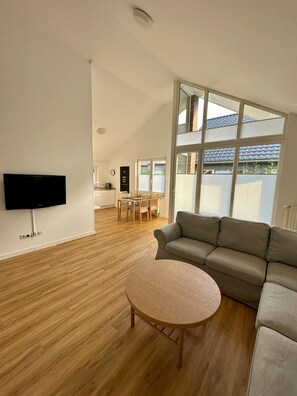 A spacious living room with a flat-screen TV, a round coffee table, a grey sofa, and a dining area with a wooden table and chairs. - Ferienhaus Fam. Mersmann (Papenburg)