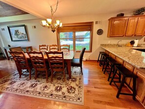 A dining area with a wooden table and chairs, a patterned rug, and a kitchen with a granite countertop and wooden cabinets.