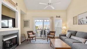 Un salon doté d’une cheminée, d’un téléviseur, d’un ventilateur de plafond, d’un canapé, de deux fauteuils, d’une table basse et offrant une vue sur un balcon aménagé avec du mobilier d’extérieur.