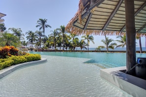 A clear swimming pool with a curved edge, surrounded by palm trees and a thatched roof structure. - Molino De Agua Beachfront 3-Bed Oasis w/ Pool, Spacious Balcony & Ocean Views (Puerto Vallarta)